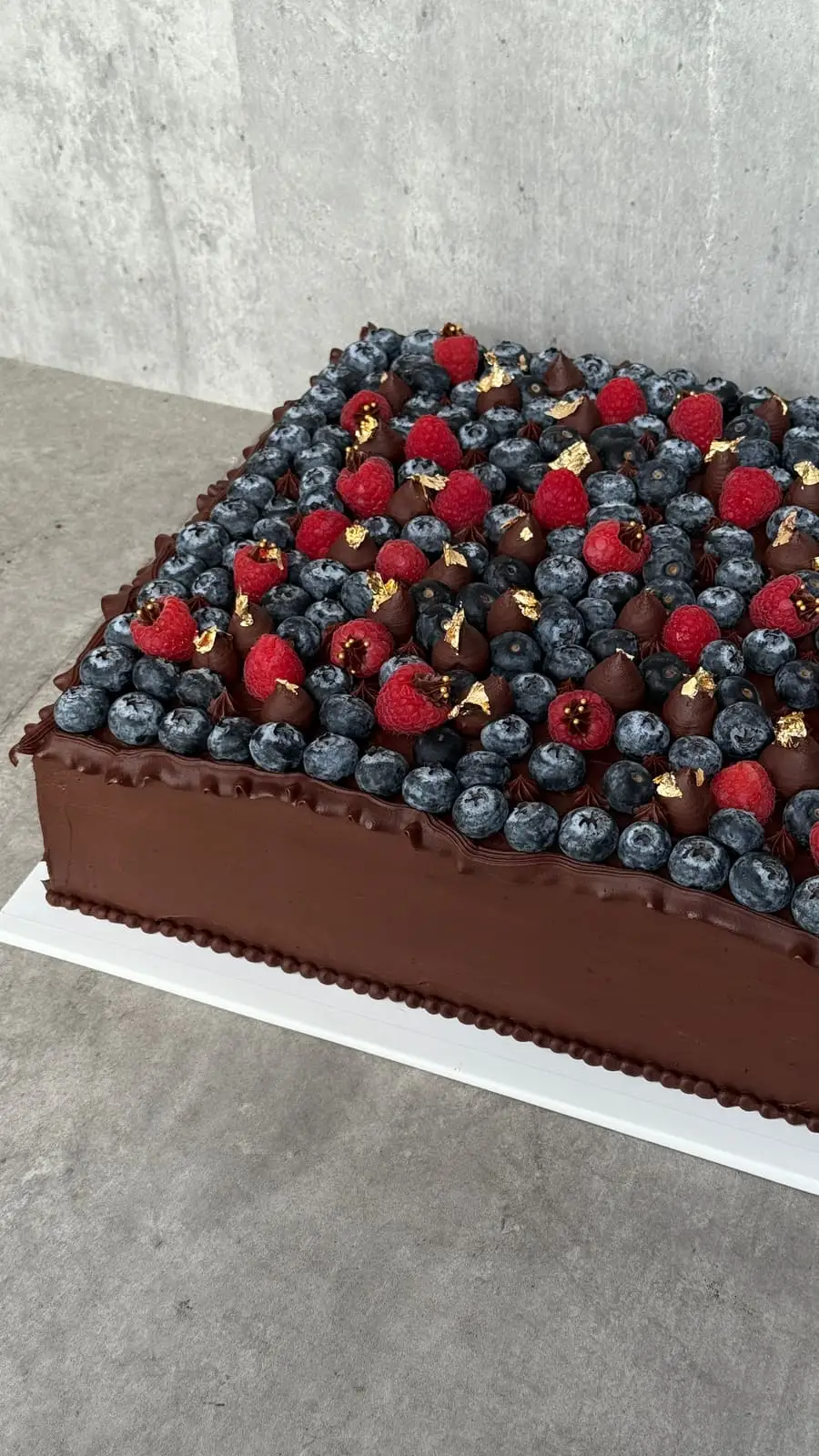 Rectangular chocolate cake covered with blueberries, raspberries, chocolate buttercream, and gold leaf.